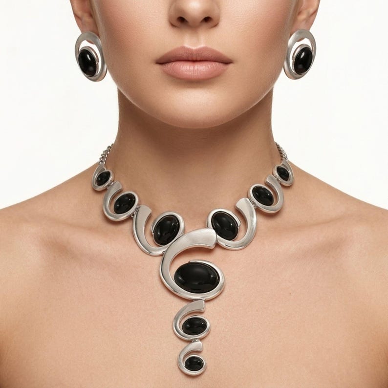Puede incluir: Un conjunto de collar y pendientes plateados con piedras negras ovaladas. El collar presenta un dise&ntilde;o &uacute;nico y fluido con elementos curvos. Los pendientes son ovalados con una piedra negra en el centro, complementando el estilo del collar.