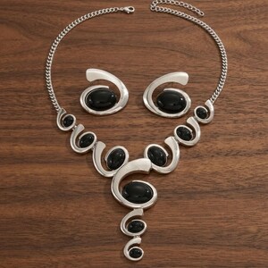 Puede incluir: Un conjunto de collar y pendientes plateados. El collar presenta una cadena y una serie de dise&ntilde;os calados en espiral, cada uno con una piedra negra ovalada grande. Los pendientes reflejan el dise&ntilde;o con una sola piedra negra.