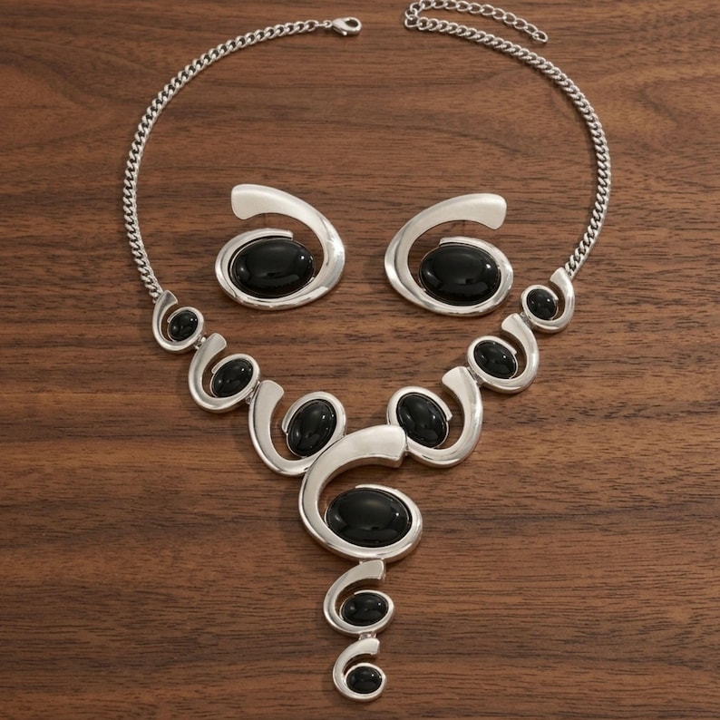 Puede incluir: Un conjunto de collar y pendientes plateados. El collar presenta una cadena y una serie de dise&ntilde;os calados en espiral, cada uno con una piedra negra ovalada grande. Los pendientes reflejan el dise&ntilde;o con una sola piedra negra.