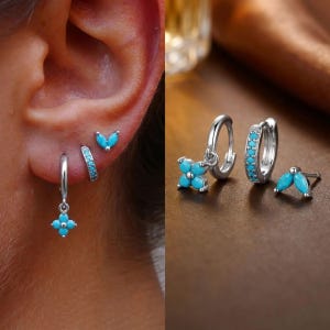 以下が含まれることがあります： ターコイズのアクセントが付いたシルバーのイヤリングのコレクション。イヤリングには、小さなスタッド、ターコイズの石が付いたフープイヤリング、ターコイズの花のデザインのイヤリングが含まれます。イヤリングは耳と茶色の表面に表示されています。