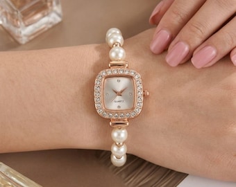 Op de afbeelding: Een roségouden quartz horloge met een vierkante wijzerplaat, omringd door kleine kristallen. De horlogeband is gemaakt van witte imitatieparels. Het horloge zit om een pols.