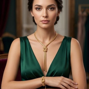 Könnte beinhalten: Eine goldfarbene Metallhalskette mit einem zentralen Knotendesign und zwei herabhängenden Strängen, kombiniert mit einem passenden goldfarbenen Metallarmband, das ebenfalls ein Knotenmotiv aufweist. Das Set wird zu einem ärmellosen dunkelgrünen Kleid getragen.