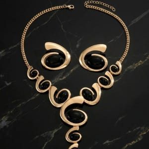 Puede incluir: Un conjunto de joyas dorado con un collar y pendientes a juego. El collar y los pendientes tienen un dise&ntilde;o en espiral con piedras negras de forma ovalada. El conjunto se exhibe sobre una superficie de m&aacute;rmol negro.