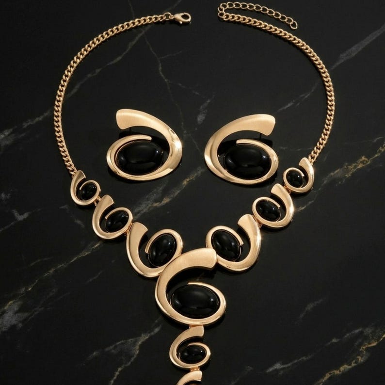 Puede incluir: Un conjunto de joyas dorado con un collar y pendientes a juego. El collar y los pendientes tienen un dise&ntilde;o en espiral con piedras negras de forma ovalada. El conjunto se exhibe sobre una superficie de m&aacute;rmol negro.
