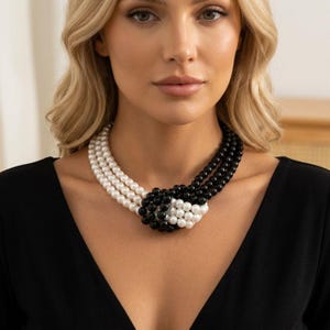 May include: A multi-strand necklace featuring alternating white and black spherical beads. The strands are gathered and intertwined at the center into a knotted design, creating a contrasting color pattern against the solid black garment.