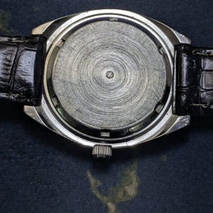 May include: Close-up of the back of a silver wristwatch with a black leather band. The watch face is a textured, circular design. The band is black and has a crocodile skin pattern.
