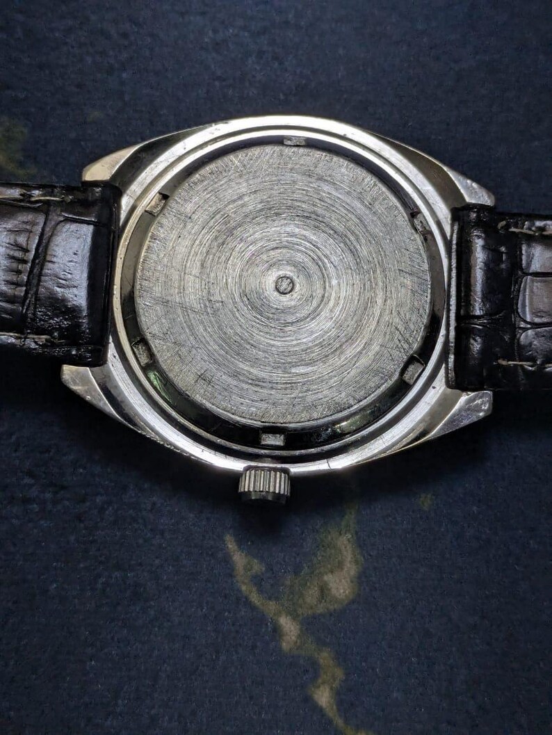 May include: Close-up of the back of a silver wristwatch with a black leather band. The watch face is a textured, circular design. The band is black and has a crocodile skin pattern.