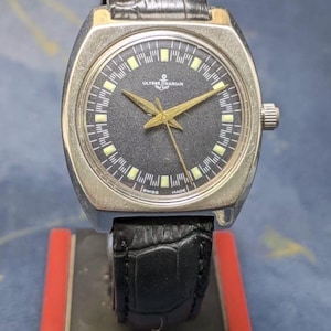 May include: A vintage Ulysse Nardin wristwatch with a black dial, gold hands, and a silver-tone case. The watch features a black leather band and is marked "Swiss Made." The watch is displayed on a red and black stand.
