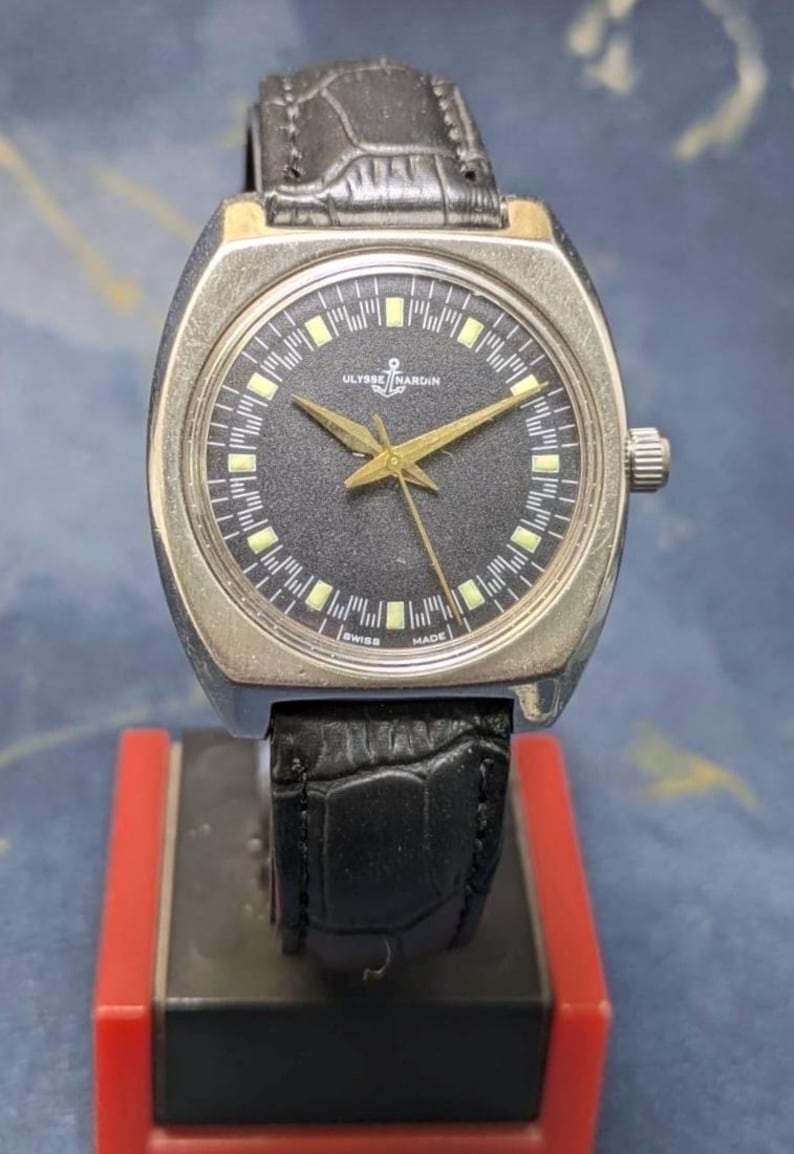 May include: A vintage Ulysse Nardin wristwatch with a black dial, gold hands, and a silver-tone case. The watch features a black leather band and is marked "Swiss Made." The watch is displayed on a red and black stand.