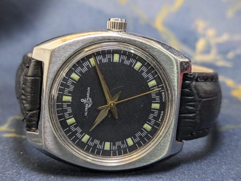 May include: A vintage Ulysse Nardin wristwatch with a black dial and silver-tone case. The watch features gold hands and markers, a black leather band, and the text "Ulysse Nardin" and "Swiss Made".