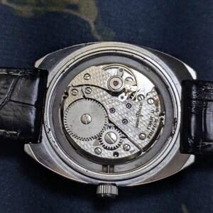 May include: Close-up of a vintage wristwatch with a transparent back, revealing the intricate mechanical movement. The watch has a silver case, a black leather band, and a small crown. The movement features gears, jewels, and the text "17 JEWELS".