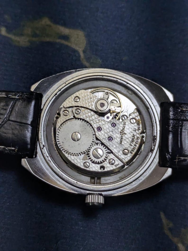 May include: Close-up of a vintage wristwatch with a transparent back, revealing the intricate mechanical movement. The watch has a silver case, a black leather band, and a small crown. The movement features gears, jewels, and the text "17 JEWELS".