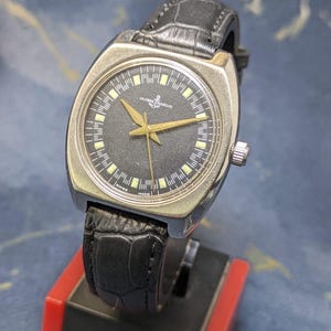 May include: A vintage Ulysse Nardin wristwatch with a black dial, gold hands, and a silver-tone case. The watch features a black leather band and is displayed on a red and black stand. The watch face has the text "Ulysse Nardin" and "Swiss Made".