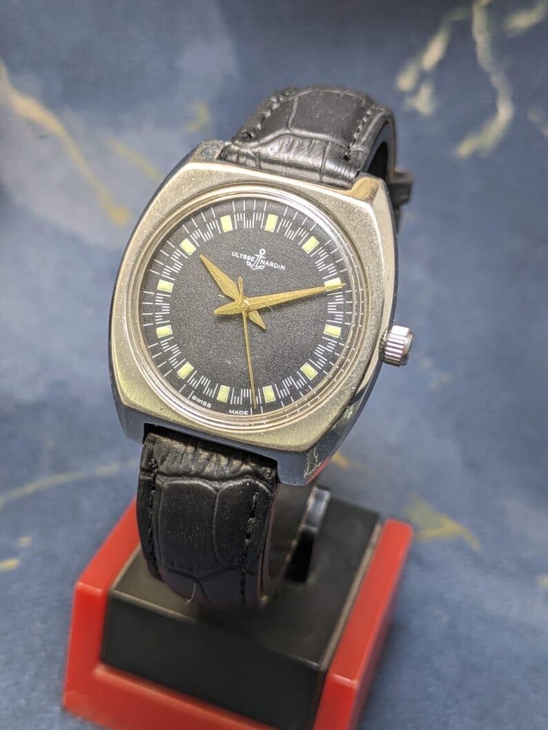 May include: A vintage Ulysse Nardin wristwatch with a black dial, gold hands, and a silver-tone case. The watch features a black leather band and is displayed on a red and black stand. The watch face has the text "Ulysse Nardin" and "Swiss Made".