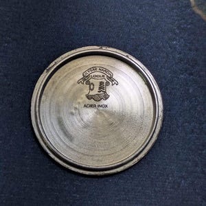 May include: A circular, metallic watch component with the text "ULYSSE NARDIN LOCLE" and "ACIER INOX" engraved on its surface. The component has a brushed finish and a raised edge.