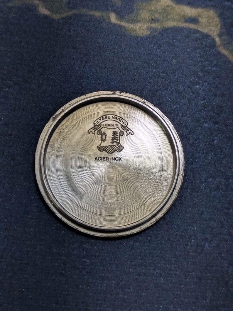May include: A circular, metallic watch component with the text "ULYSSE NARDIN LOCLE" and "ACIER INOX" engraved on its surface. The component has a brushed finish and a raised edge.