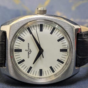 May include: A silver Ulysse Nardin watch with a white face and black markers. The watch has a black leather band. The watch face displays the Ulysse Nardin logo and the words "MADE-T SWISS".