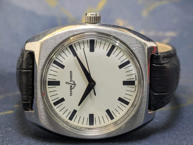 May include: A silver Ulysse Nardin watch with a white face and black markers. The watch has a black leather band. The watch face displays the Ulysse Nardin logo and the words "MADE-T SWISS".