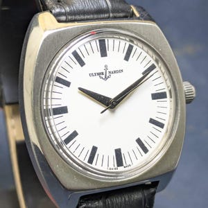 May include: A silver-toned Ulysse Nardin watch with a square face and a black leather band. The white watch face features black hour markers and hands. The watch has the text "ULYSSE NARDIN" and "T-SWISS MADE-T" printed on the face.