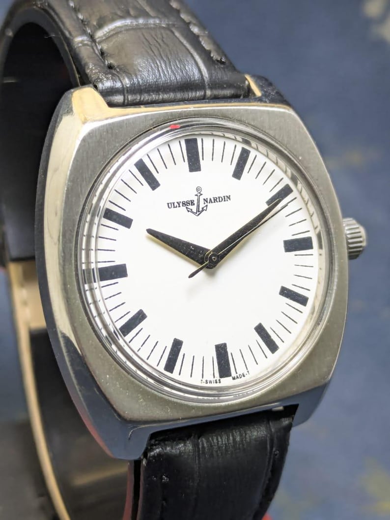 May include: A silver-toned Ulysse Nardin watch with a square face and a black leather band. The white watch face features black hour markers and hands. The watch has the text "ULYSSE NARDIN" and "T-SWISS MADE-T" printed on the face.