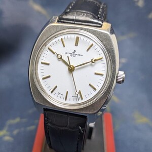 May include: A vintage Ulysse Nardin wristwatch with a silver-toned case and a black leather band. The watch features a white face with gold-colored hands and markers. The watch is marked "Swiss Made".