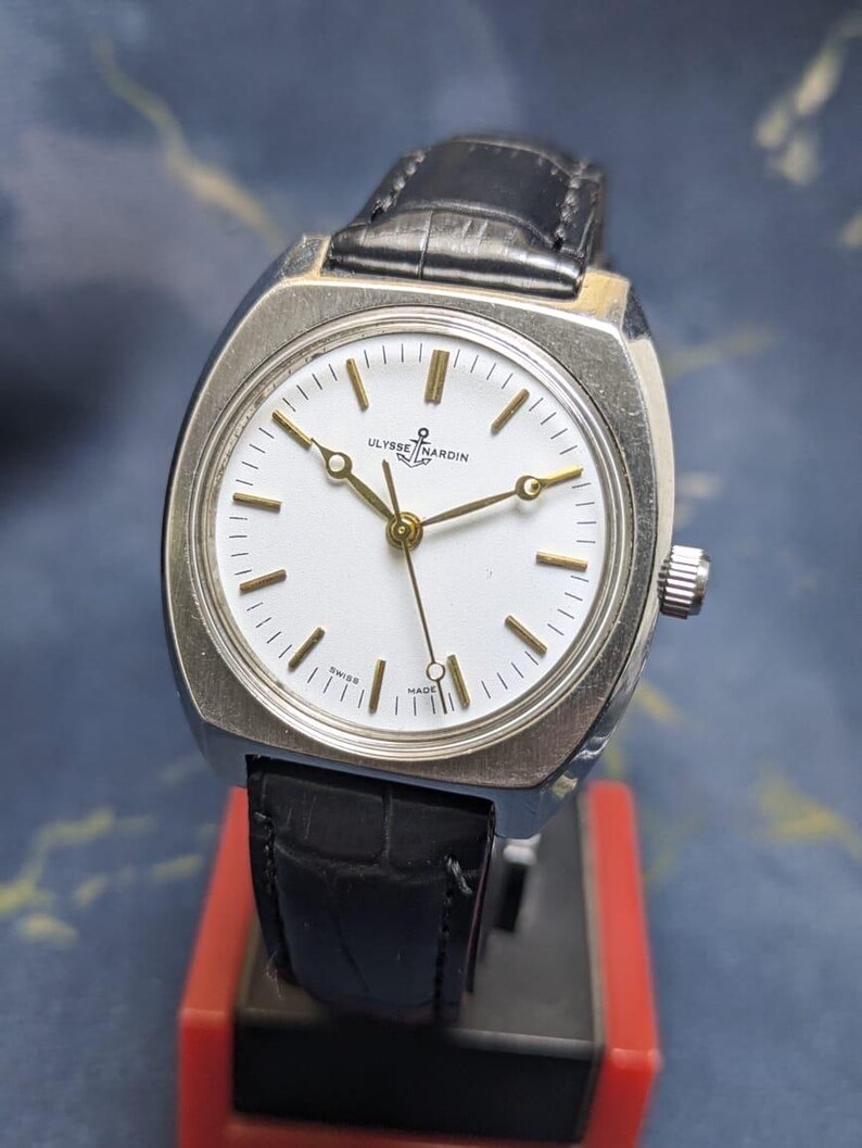 May include: A vintage Ulysse Nardin wristwatch with a silver-toned case and a black leather band. The watch features a white face with gold-colored hands and markers. The watch is marked "Swiss Made".