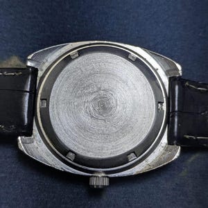 May include: Close-up of the back of a vintage wristwatch. The watch has a silver-colored metal case and a black leather strap. The watch face is a textured, circular design. The watch's crown is visible on the side.