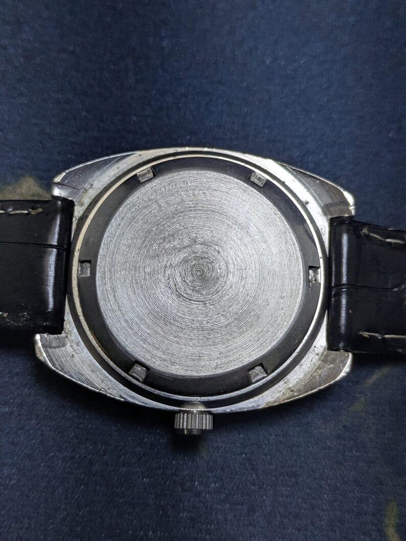 May include: Close-up of the back of a vintage wristwatch. The watch has a silver-colored metal case and a black leather strap. The watch face is a textured, circular design. The watch's crown is visible on the side.