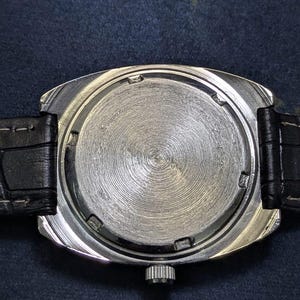 May include: A close-up of the back of a silver-toned wristwatch with a black leather band. The watch face has a circular, textured design. The band is black and appears to be made of leather. The watch has a rectangular shape.