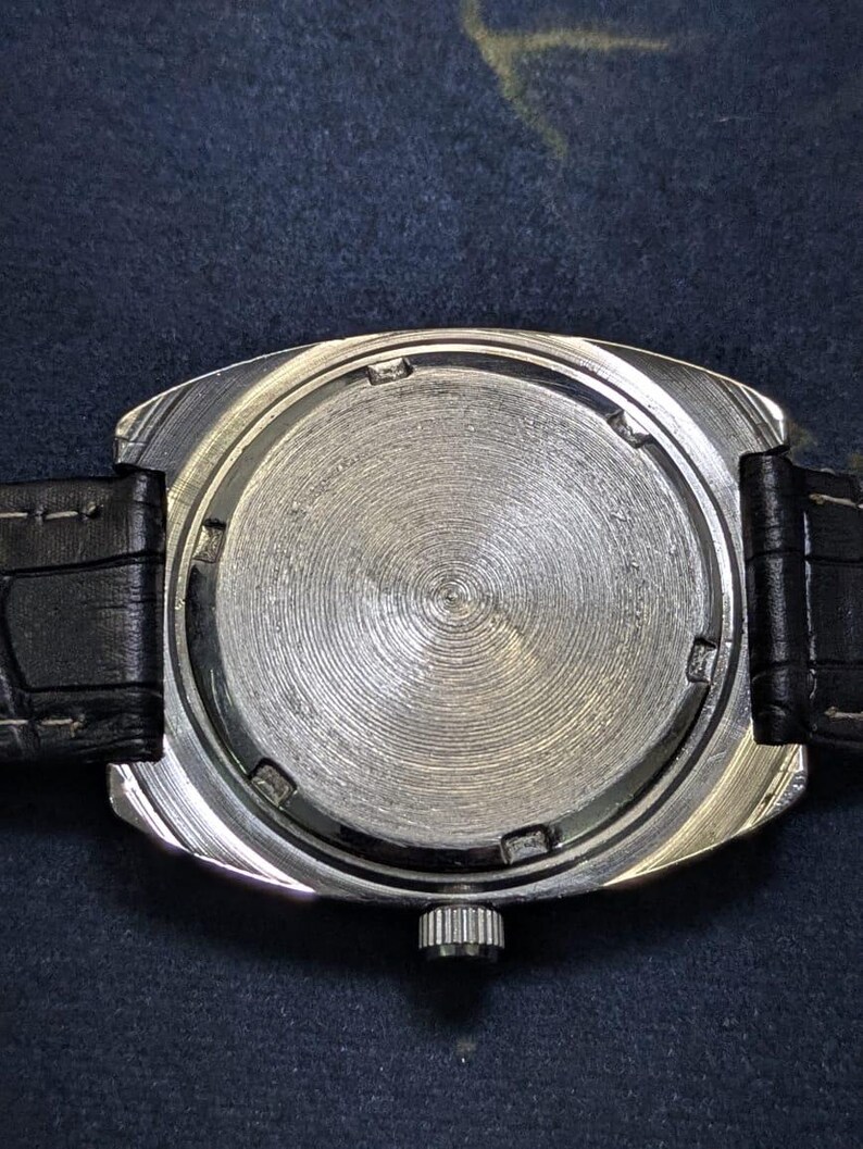 May include: A close-up of the back of a silver-toned wristwatch with a black leather band. The watch face has a circular, textured design. The band is black and appears to be made of leather. The watch has a rectangular shape.