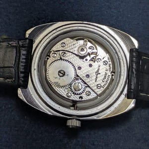 May include: Close-up of a vintage wristwatch with the back cover removed, revealing the intricate mechanical movement. The watch has a silver-toned case and a black leather strap. The movement features gears, jewels, and the text "DUTCH MARIN" and "SWISS SEVENTEEN JEWELS".