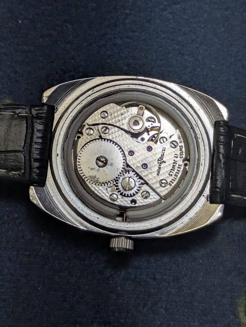 May include: Close-up of a vintage wristwatch with the back cover removed, revealing the intricate mechanical movement. The watch has a silver-toned case and a black leather strap. The movement features gears, jewels, and the text "DUTCH MARIN" and "SWISS SEVENTEEN JEWELS".