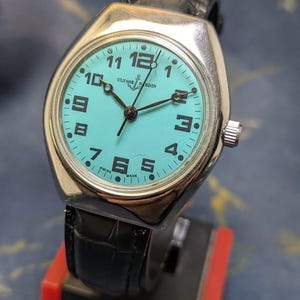 May include: A vintage Ulysse Nardin wristwatch with a silver case and a turquoise dial. The watch features black numerals and hands, a black leather band, and a crown on the side. The watch is displayed on a red and black stand.