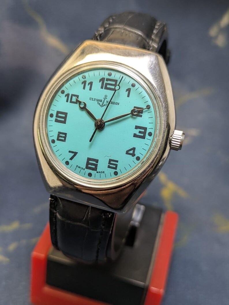 May include: A vintage Ulysse Nardin wristwatch with a silver case and a turquoise dial. The watch features black numerals and hands, a black leather band, and a crown on the side. The watch is displayed on a red and black stand.