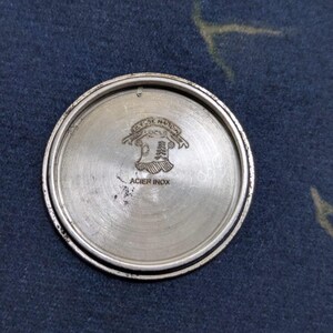 May include: A silver-colored watch back with the text "Ulysse Nardin Locle" and "Acier Inox" etched into the metal. The watch back has a circular shape with a raised outer rim and a brushed metal finish.
