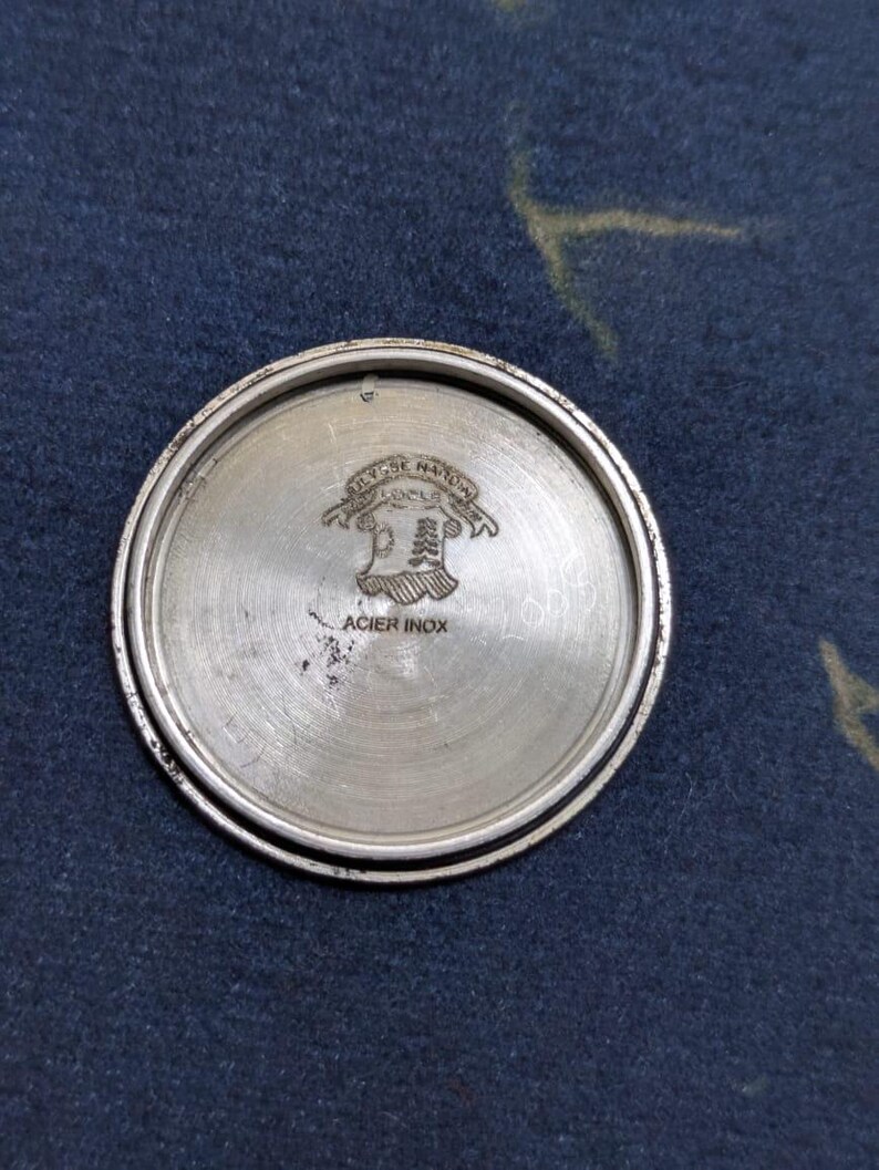 May include: A silver-colored watch back with the text "Ulysse Nardin Locle" and "Acier Inox" etched into the metal. The watch back has a circular shape with a raised outer rim and a brushed metal finish.