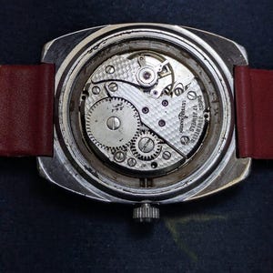 May include: A detailed view of a vintage Ulysse Nardin watch, revealing the internal workings. The watch has a silver case, a red leather strap, and a crown. The mechanism displays gears, jewels, and the inscription "Ulysse Nardin 17 Jewels Swiss".