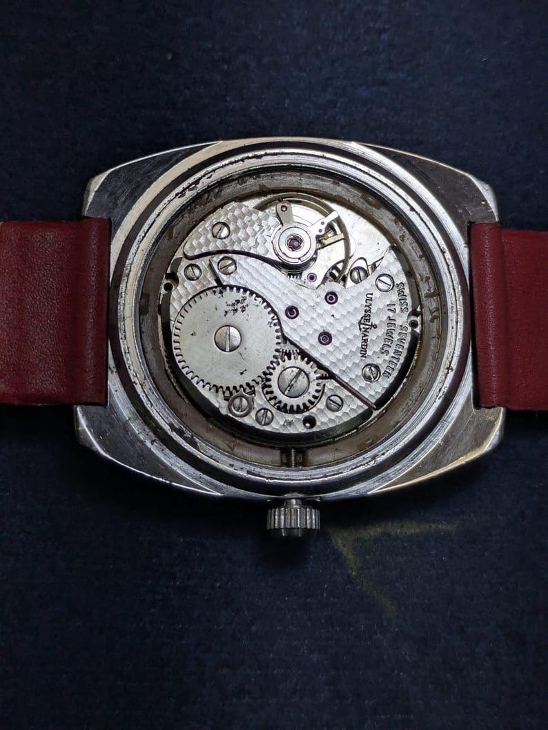 May include: A detailed view of a vintage Ulysse Nardin watch, revealing the internal workings. The watch has a silver case, a red leather strap, and a crown. The mechanism displays gears, jewels, and the inscription "Ulysse Nardin 17 Jewels Swiss".