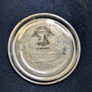 May include: A silver-toned watch back with the engraved text "ULYSSE NARDIN LOCLE" above a crest design. The words "ACIER INOX" are also visible. The watch back is circular and has a brushed metal finish.