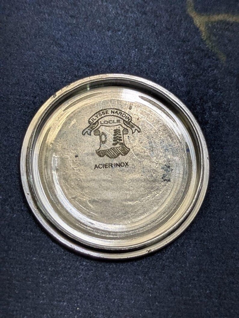May include: A silver-toned watch back with the engraved text "ULYSSE NARDIN LOCLE" above a crest design. The words "ACIER INOX" are also visible. The watch back is circular and has a brushed metal finish.
