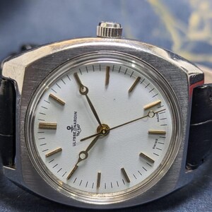 May include: A vintage Ulysse Nardin wristwatch with a silver-toned case and a white dial. The watch features gold-colored hands and markers, and a black leather band. The dial is marked "Swiss Made".