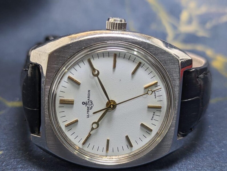 May include: A vintage Ulysse Nardin wristwatch with a silver-toned case and a white dial. The watch features gold-colored hands and markers, and a black leather band. The dial is marked "Swiss Made".