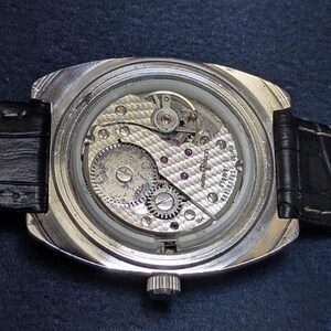 May include: Close-up of a vintage wristwatch, showcasing the intricate mechanical movement. The watch features a silver-toned case, a black leather strap, and a crown. The internal gears, jewels, and other components are visible through the clear back.
