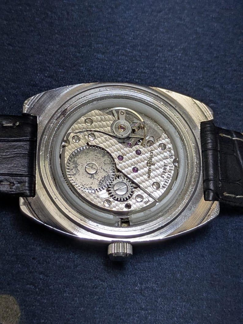 May include: Close-up of a vintage wristwatch, showcasing the intricate mechanical movement. The watch features a silver-toned case, a black leather strap, and a crown. The internal gears, jewels, and other components are visible through the clear back.