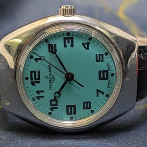 May include: A vintage Ulysse Nardin watch with a teal face and black numerals. The watch has a silver-toned case and a black leather band. The watch face displays the brand name and "Swiss Made".