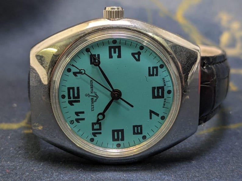 May include: A vintage Ulysse Nardin watch with a teal face and black numerals. The watch has a silver-toned case and a black leather band. The watch face displays the brand name and "Swiss Made".