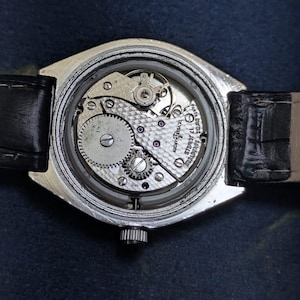 May include: A silver-toned wristwatch with a black leather band. The watch's back is open, revealing the intricate internal mechanism, including gears, jewels, and the inscription "Ulysse Nardin 17 Jewels Swiss".
