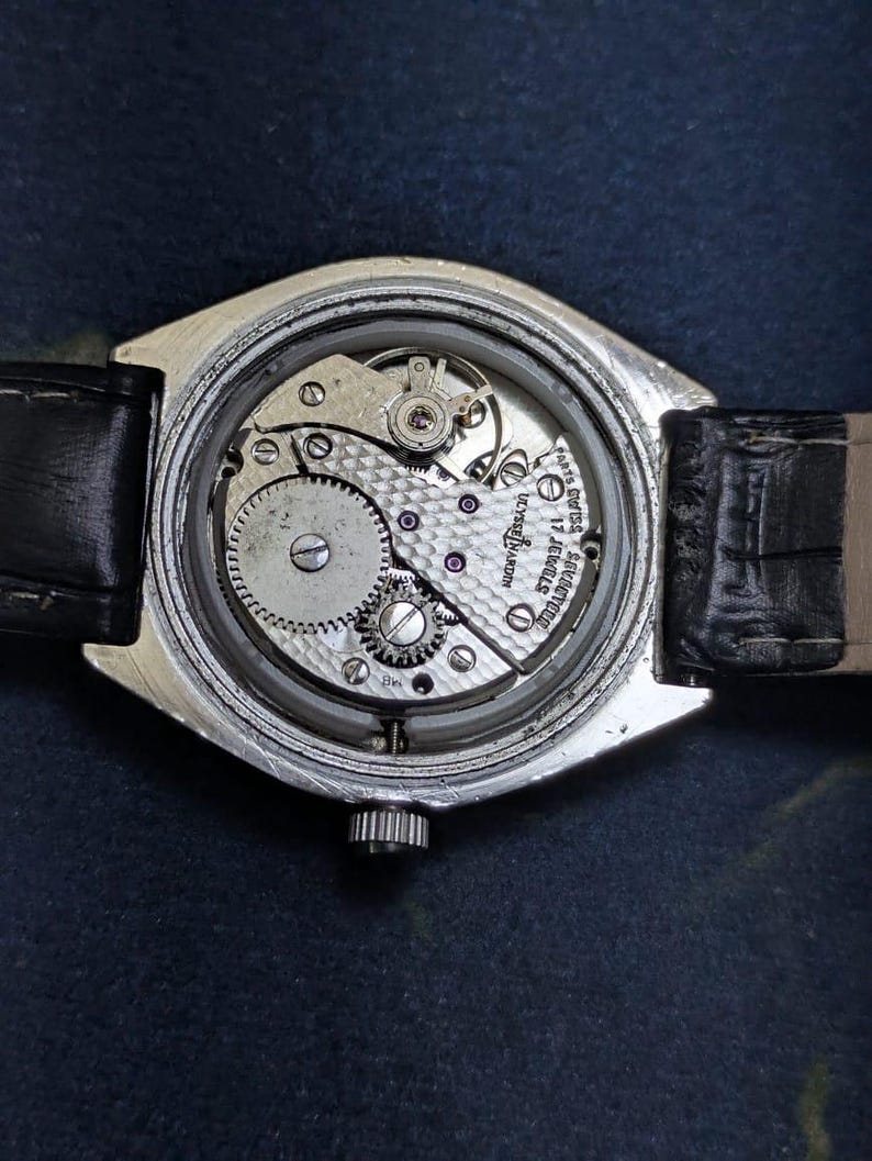 May include: A silver-toned wristwatch with a black leather band. The watch's back is open, revealing the intricate internal mechanism, including gears, jewels, and the inscription "Ulysse Nardin 17 Jewels Swiss".