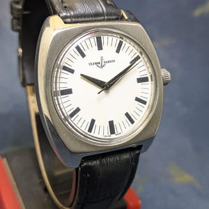 May include: A silver-toned wristwatch with a white face and black hands and markers. The watch features the brand name "Ulysse Nardin" and a black leather band. The watch has a square face with rounded corners and a small crown on the side.