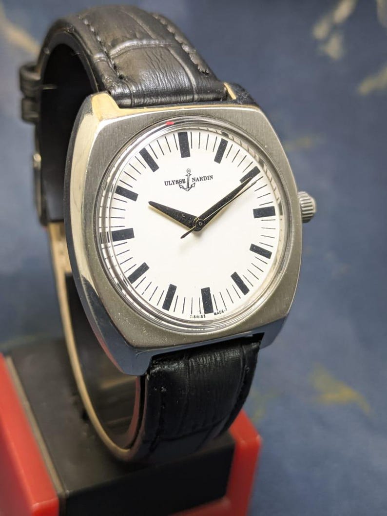 May include: A silver-toned wristwatch with a white face and black hands and markers. The watch features the brand name "Ulysse Nardin" and a black leather band. The watch has a square face with rounded corners and a small crown on the side.